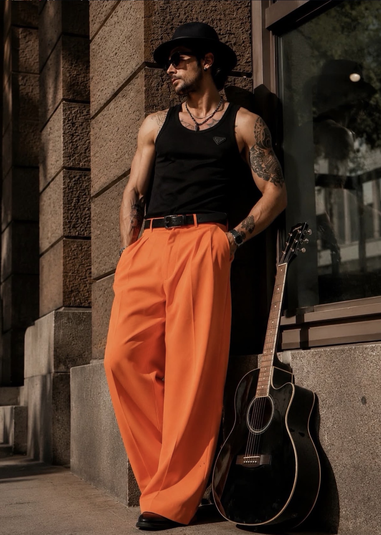 Anil in a black tank top and wide orange trousers with a guitar beside a stone facade