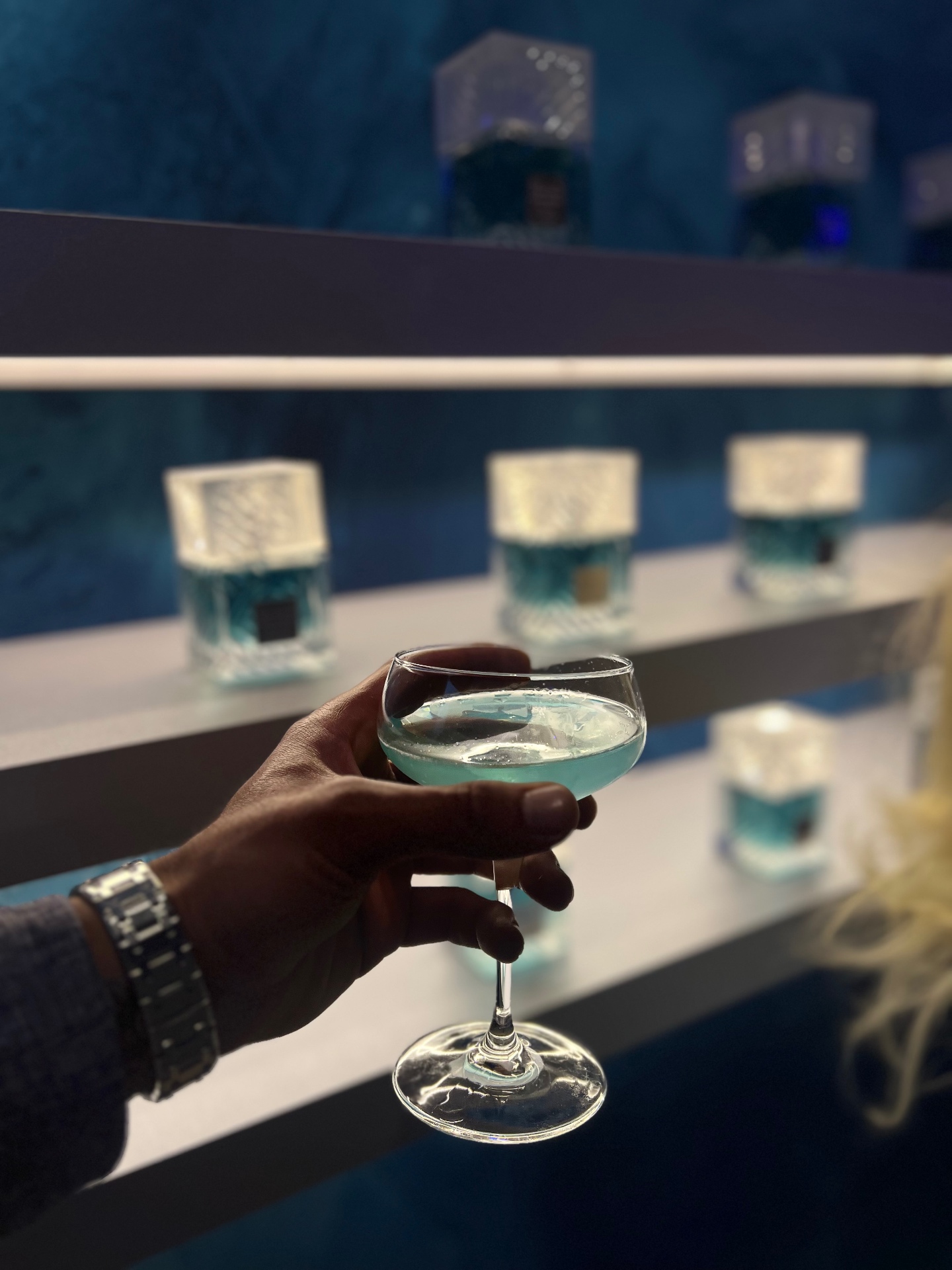 Anil's hand holding a blue cocktail in a coupe glass with fragrance bottles in the background
