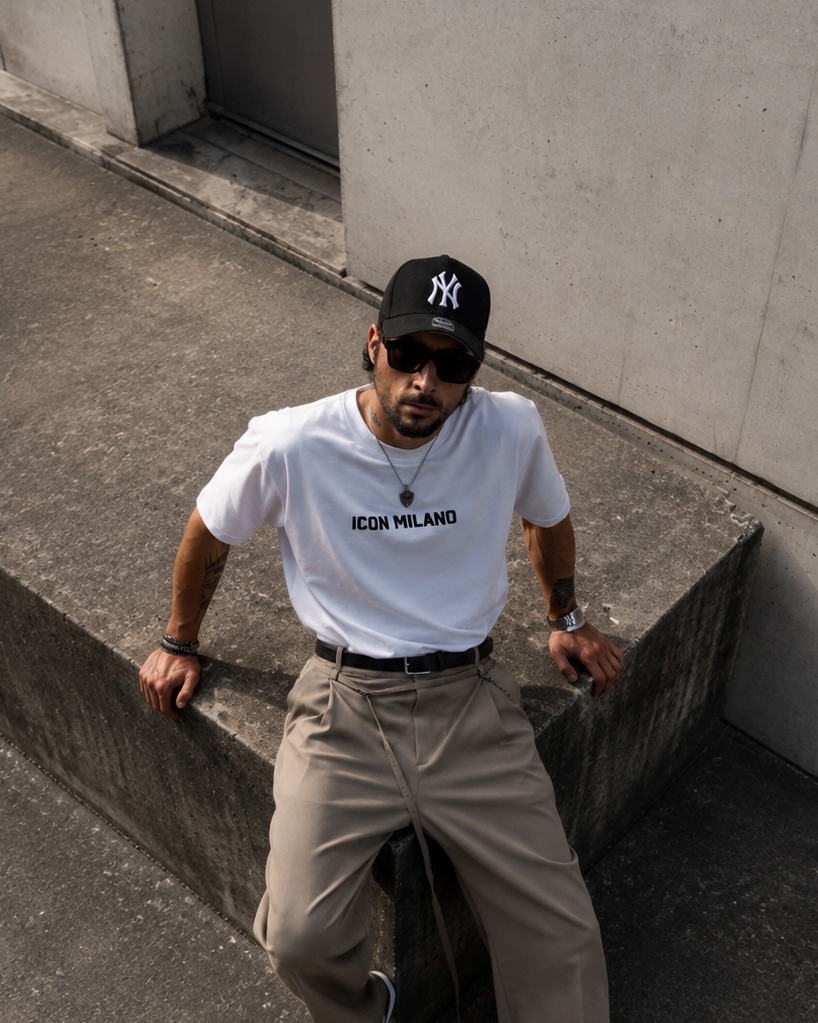 Anil in a white logo tee and taupe trousers seated against concrete architecture
